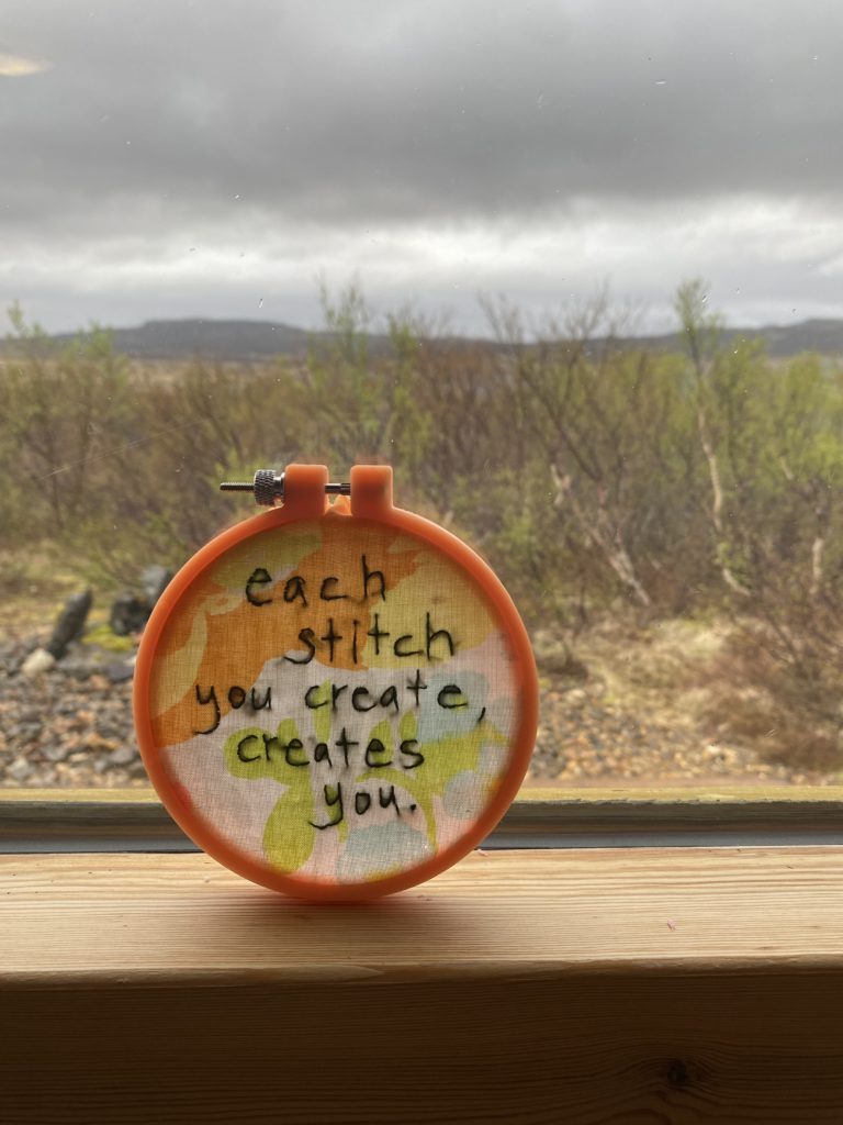 An orange embroidery hoop holds a vintage piece of floral fabric onto which has been stitched "Each stitch you create, creates you." Behind the frame are low hills, shrubs and a great sky
